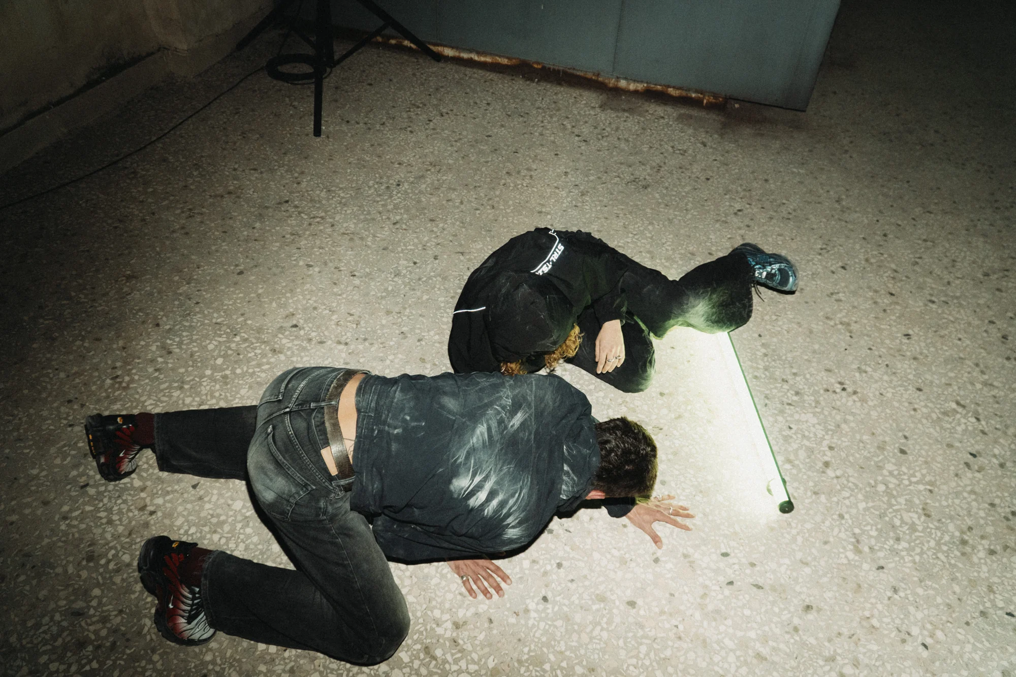 Two performers curl towards a light tube on the floor.