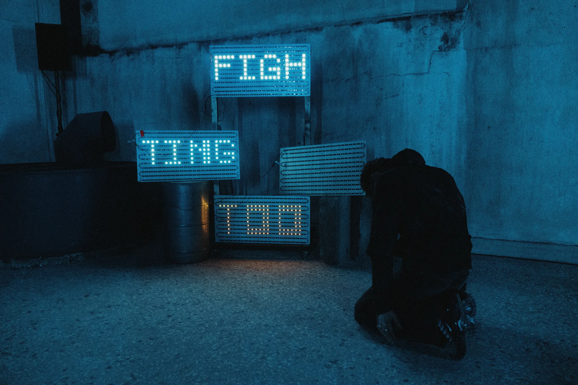 A performer crouches beside an arrangement of pixelated LED text in blue light.