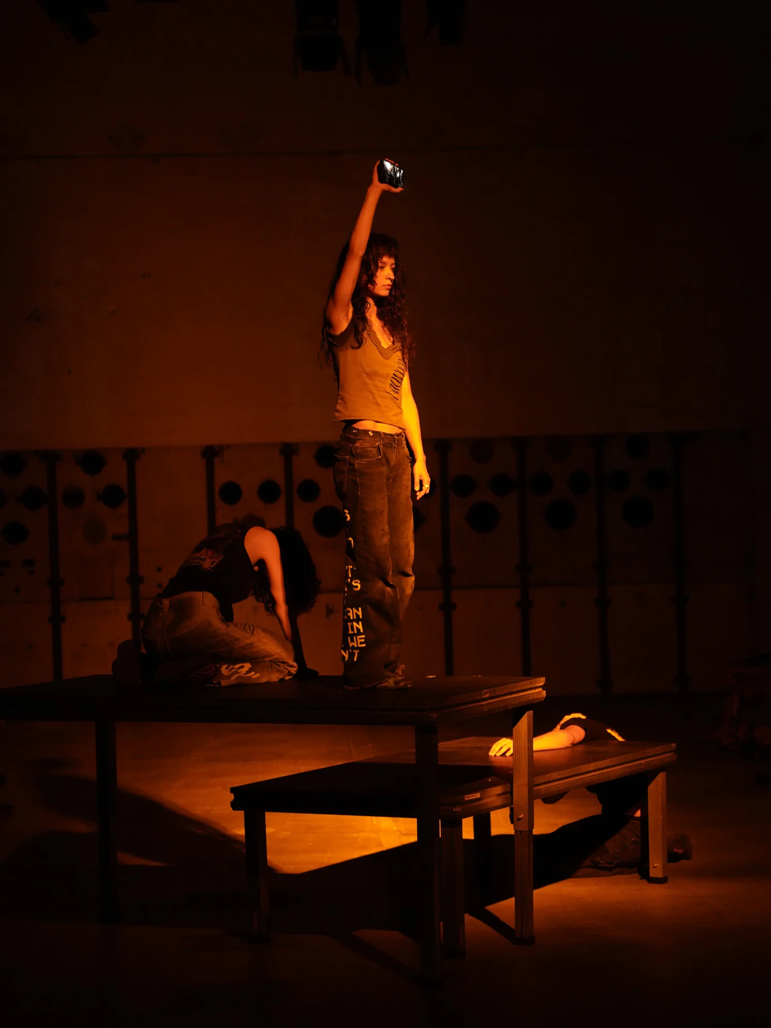 A performer raises a phone above two others on the platforms in amber light.