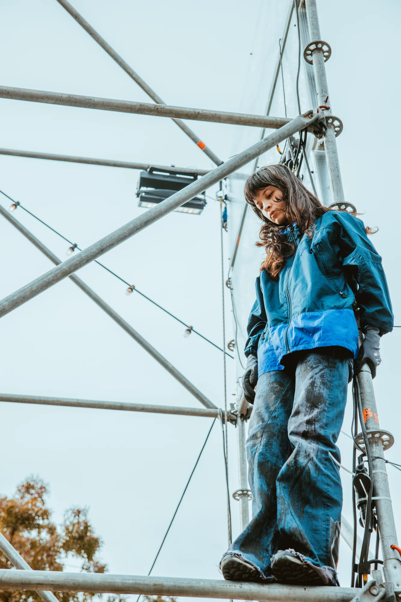 A performer stands high on the outdoor scaffolding in daylight.