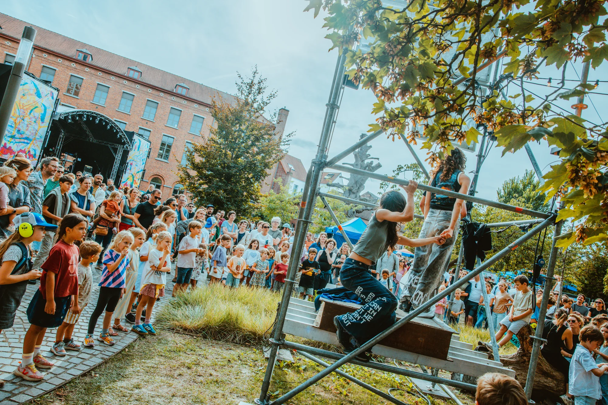 Wide view of performers on scaffolding in a public park, with the festival audience gathered beside them.